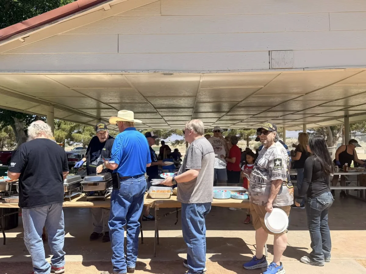 DAV Chapter 12 volunteers serve food to Veterans and families at an outdoor picnic under a pavilion, with a line of attendees holding plates.