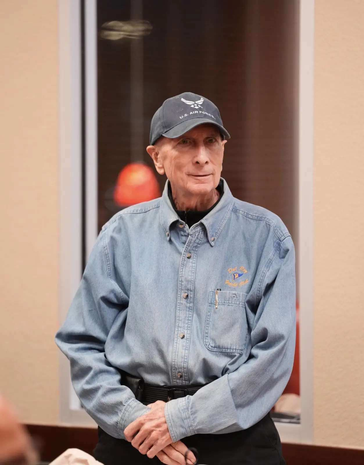 An older Veteran in a denim shirt and cap stands to address the room.