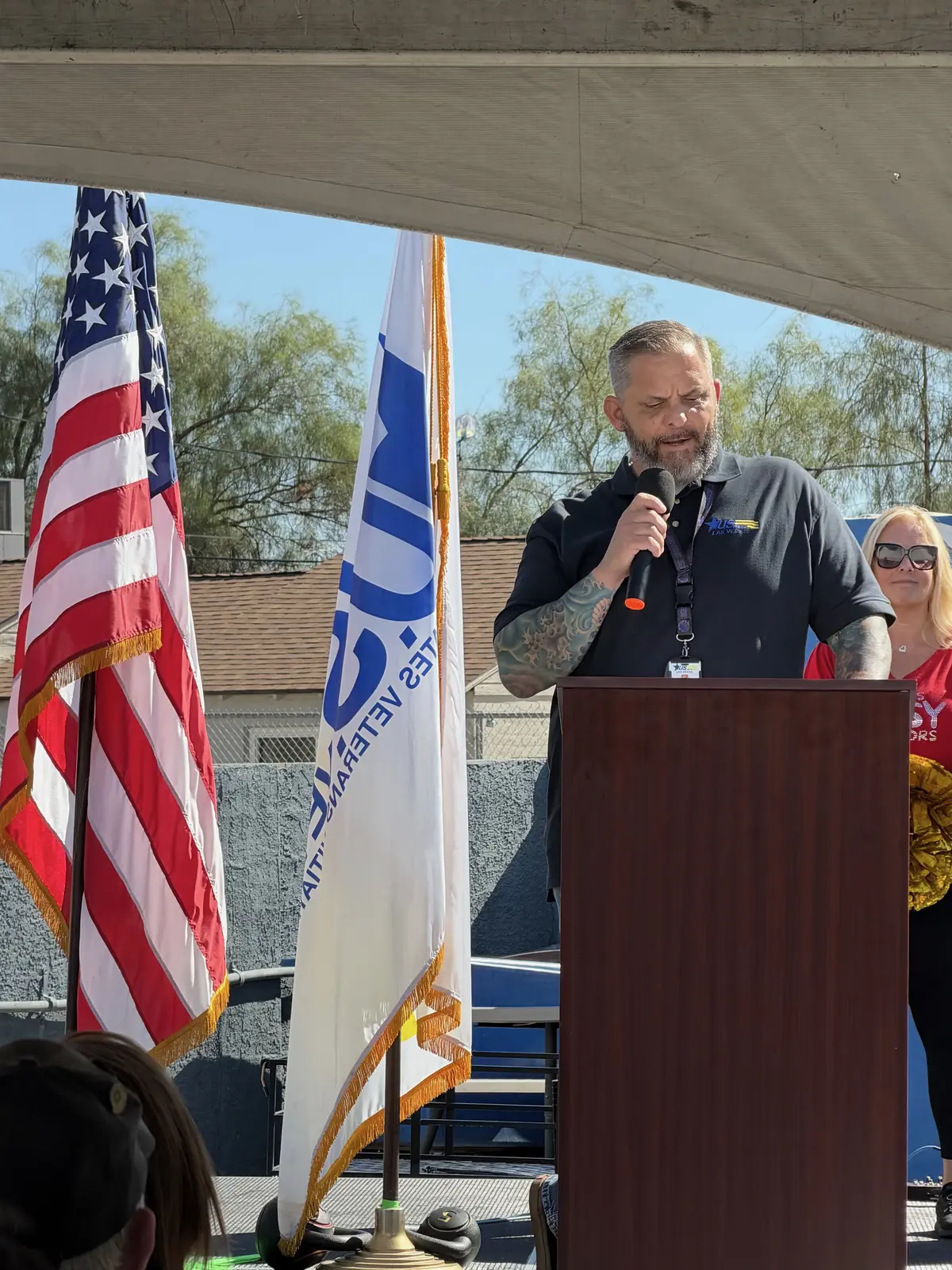 A speaker addresses the crowd from the podium, flanked by the American flag and the U.S.VETS flag.