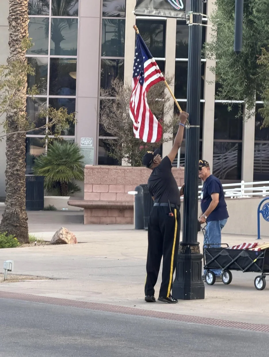 An Honor Guard member in uniform trousers with a gold stripe reaches up to remove a flag from a light pole while another member waits with a wagon.