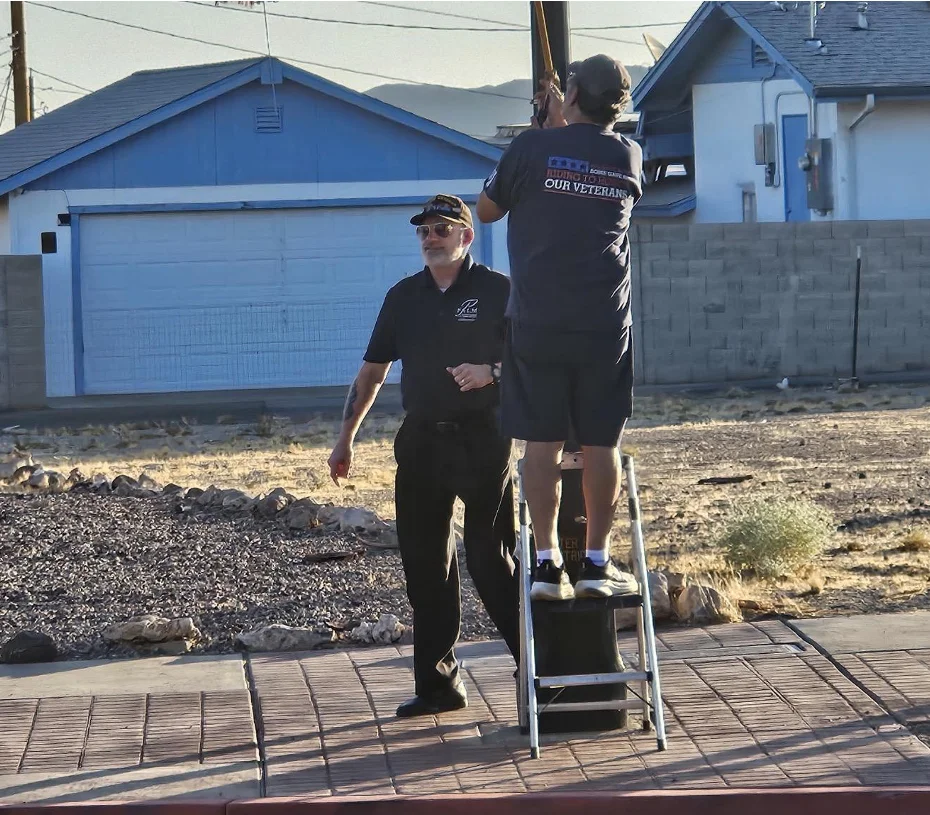 A member on a step ladder reaches a flag high on a pole as an Honor Guard member stands beside him.