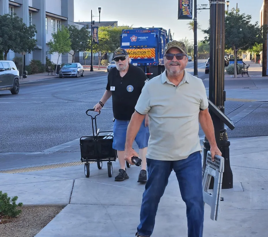 A smiling Veteran in a Marines cap carries a folding step stool while another member pulls a wagon along the sidewalk.